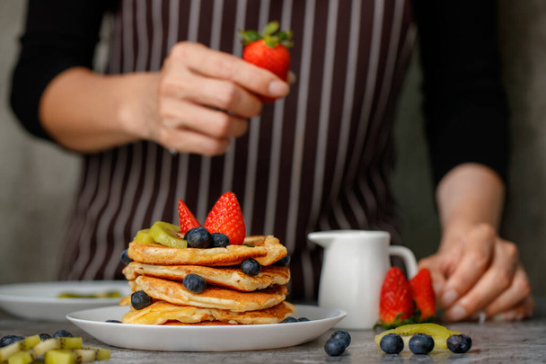 Woman housewife preparing pancakes to be food for family, putting topping fruit, strawberries, kiwi and blueberries on top. - Photo, Image