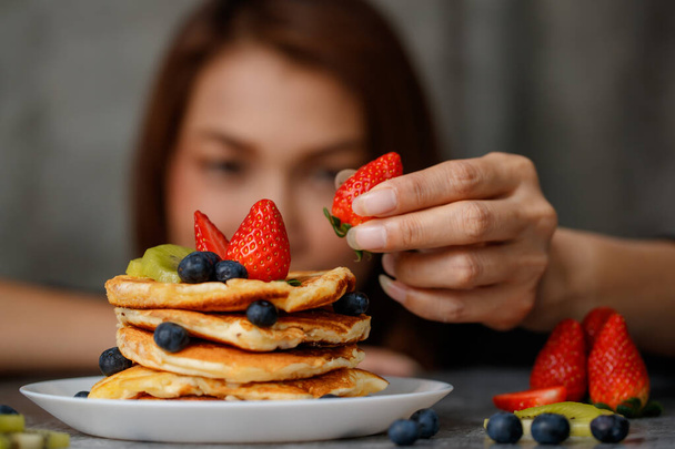 Asian housewife preparing pancakes for family, putting topping, strawberries, blueberris and kiwi on top. - Фото, изображение