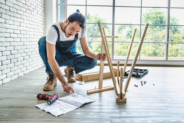 Hipster jongeman werkzaam als klusjesman op zoek naar handmatige tijdens het monteren van houten tafel met apparatuur, concept voor thuis diy en zelfbediening.in het kantoor. - Foto, afbeelding