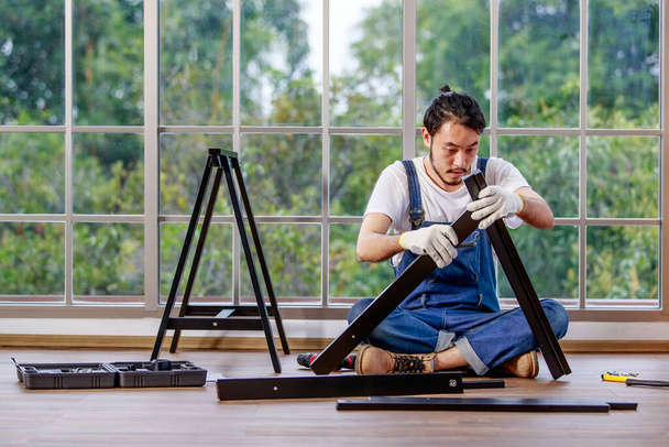 Junger asiatischer Hipster mit Bart arbeitet als Heimwerker, montiert Holztisch mit Geräten, Konzept für Heimwerker, Hobby und Selbstbedienung, grüner Baum im Hintergrund - Foto, Bild