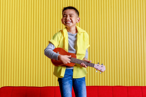 Lovely cutout portrait of young Asian boy on grey sweater and yellow shirt smile and happy for funny dance by raising hands up with small guitar as champion winner of musician and song contest - Photo, Image