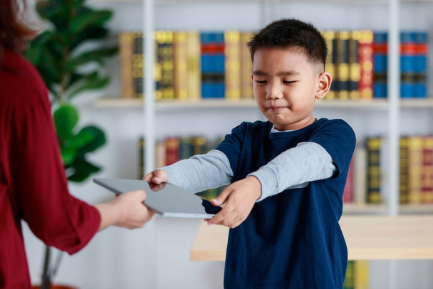 Χαρούμενο νεαρό ασιατικό αγόρι σε γκρι πουλόβερ και denim μπλε πουκάμισο στέκεται στη βιβλιοθήκη με χαρά και χαμόγελο, ενώ απλώνει τα χέρια έξω στο κόκκινο πανί πρόσωπο για να πάρει tablet για τη μελέτη στοιχειώδη γνώση - Φωτογραφία, εικόνα