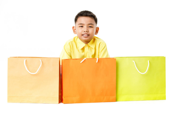 Вырезанный портрет маленького симпатичного азиатского мальчика на желтой рубашке улыбается и сидит за 3 привлекательными бумажными пакетами коричневого, оранжевого, зеленого цвета для грандиозной распродажи детскому клиенту в универмаге - Фото, изображение