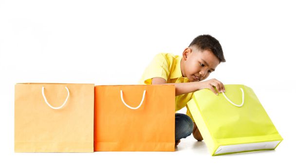 Cutout portrait of little cute Asian boy on yellow shirt smile and sit behind 3 attractive shopping paper bag of brown, orange, green for grand sale promotion to kid customer at department store - Φωτογραφία, εικόνα