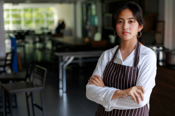 Confident ethnic female waitress in apron standing with crossed arms in cafe and looking at camera. - Фото, изображение