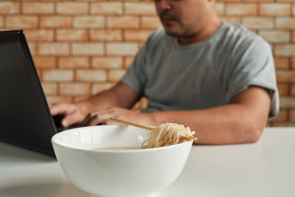 タイの男性労働者のラップトップでの作業に忙しく、急いでオフィスランチの休憩中にインスタントラーメンを食べるために箸を使用するため、迅速、おいしい、と安い。アジアのファーストフード、不健康なライフスタイル. - 写真・画像
