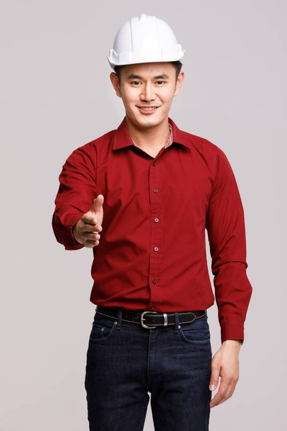 Delighted Asian male contractor in hardhat reaching out hand for greeting while standing on gray background in studio and looking at camera - Photo, Image