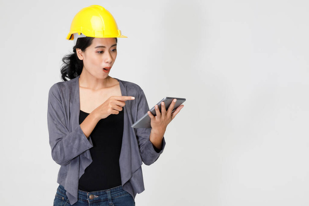 Delighted aziatische vrouwelijke ingenieur in helm staan in studio en browsing tablet op grijze achtergrond met verrassing en opgewonden gezicht. - Foto, afbeelding