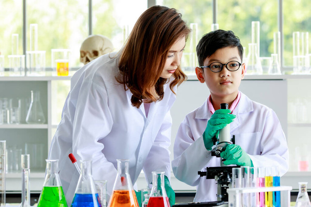 Сконцентрований азіатський хлопчик вивчає рідини через мікроскоп під час проведення експерименту в шкільній лабораторії з вчителькою-жінкою. - Фото, зображення