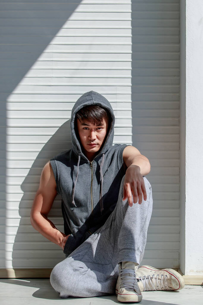 Retrato de asiático bonito homem sentado de mãos dadas no bolso camisa vestindo mangas t-shirt cinza cobrir a cabeça sportswear no fundo - Foto, Imagem