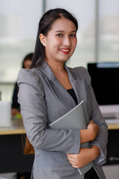 Porträt einer jungen asiatischen Geschäftsfrau, die im modernen Büro Aktenblätter hält und in die Kamera lächelt. - Foto, Bild