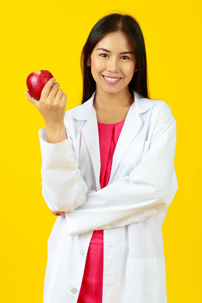 Asiatico giovane bella gentile medico femminile indossa uniforme abito bianco con stetoscopio, sorridente e guardando mela rossa. Presenta nutrizione, vitamina, sano con sfondo giallo isolato - Foto, immagini