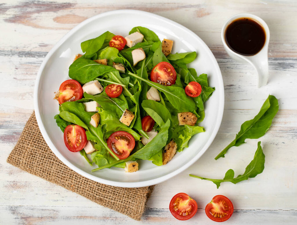 Fresh spinach salad with tomato, small pieces of herb roasted chicken in white ceramic dish on brown sackcloth, together with balsamic vinegar on white wooded table. Concept for healthy. - Photo, Image