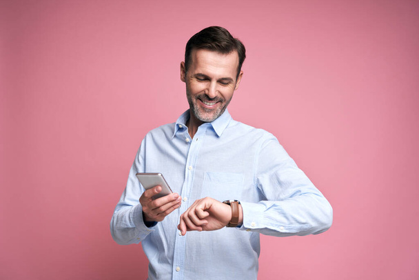 Studio shot of middle aged caucasian man using phone and checking time on watch - Foto, Imagen