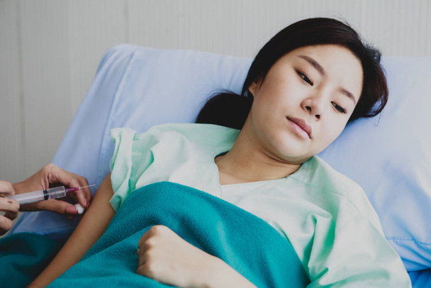 Aasialainen nuori pitkäkarvainen naispotilas pukeutuu vihreään sairaalan univormuun, joka on peitetty peitolla, makaa sinisellä tyynysängyllä tuntea kipua, kun lääkäri ampui lääkkeen injektiota neulalla ja ruiskulla oikeaan käsivarteen.. - Valokuva, kuva