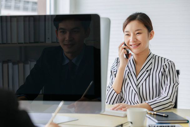 アジアの美しい女性従業員で黒と白のストライプシャツに座っています椅子笑顔話上の電話一方で男性労働者上の反対側のオフィスデスク誰影を反映上のコンピュータモニタpeek上の彼女の. - 写真・画像