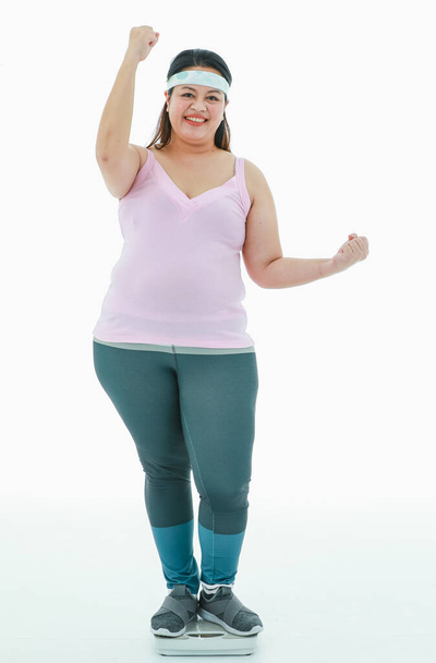 Portrait isolé plan de studio d'Asiatique heureux forte fille de graisse réussie dans les leggings de vêtements de sport avec bandeau debout sur l'échelle de poids tenir le poing en l'air après avoir reçu la victoire dans la perte de poids. - Photo, image