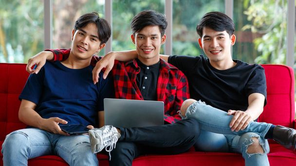 Счастливые молодые азиаты с планшетом и ноутбуком улыбаются и смотрят в камеру, сидя дома на диване - Фото, изображение