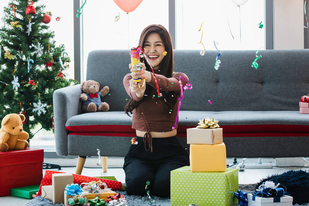 Asian happy beautiful female sitting on carpet surrounded by colorful present gift boxes and teddy bear dolls smiling laughing when pulling party popper paper confetti celebrating Christmas festival. - Photo, image