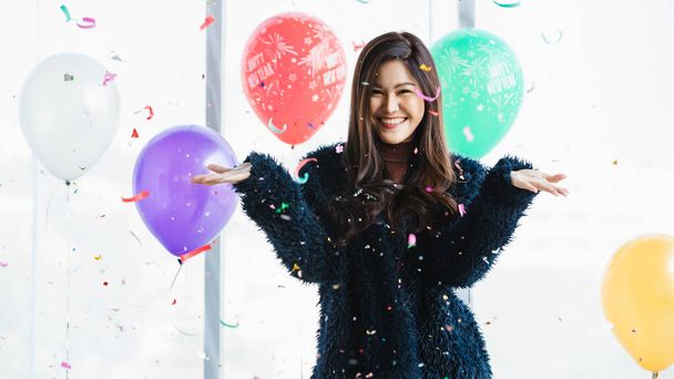 Aasian nuori onnellinen kaunis nainen sininen karvainen takki seisoo hymyillen katso kameraa vaaleanpunainen paperi popper confetti edessä värikäs uudenvuoden juhla koristelu ilmapalloja tausta. - Valokuva, kuva