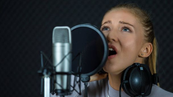 Młoda piosenkarka wykonująca piosenkę w pobliżu mikrofonu w profesjonalnym studiu nagraniowym z dźwiękoszczelną ścianą - Zdjęcie, obraz