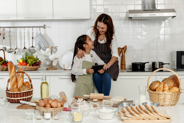 Asian mother and cute daughter, happy young mother teach cook together with small daughter doing bakery and learning computer and laptop to make cake in the kitchen - Zdjęcie, obraz