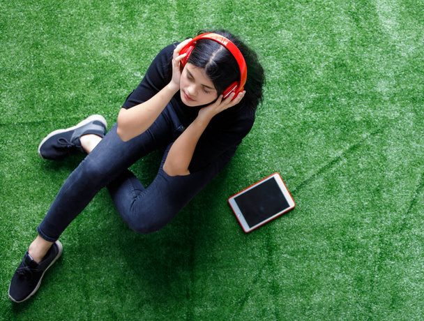 Bird-eye view portrait shot of an attractive young teenager enjoying listening to music with tablet and headset. Junior girl with casual clothes sitting and relaxing on the artificial grass - Fotografie, Obrázek