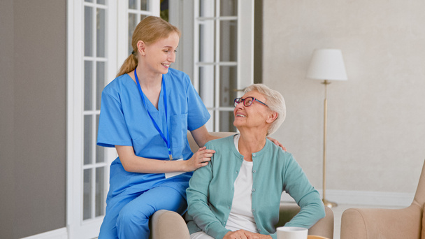 Молодая медсестра в форме слушает старшего пациента, сидя на подлокотнике удобного кресла - Фото, изображение