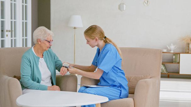 Skilled nurse in blue uniform puts digital blood pressure monitor on senior woman hand in light office - Photo, Image