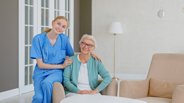 Nuori blondi sairaanhoitaja univormussa huolehtii vanhempi nainen valoisa huone asumiseen - Valokuva, kuva