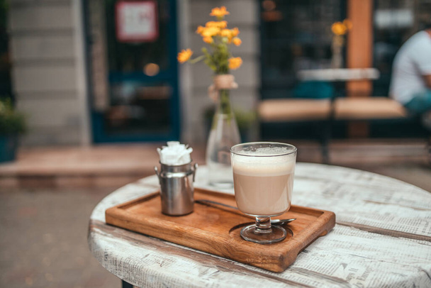 外の通りのカフェで居心地の良いヴィンテージテラスの木製テーブルの上の香りのカプチーノコーヒーカップ - 写真・画像