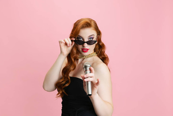 レトロな髪型とピンクの背景に孤立マイクで流行のサングラスの歌と効果的なスタイリッシュな女性の肖像画 - 写真・画像