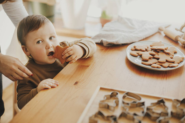Cute little girl eating freshly baked gingerbread cookie on wooden table in modern room. Authentic lovely moment, holiday preparations. Adorable funny toddler tasting christmas cookies - Photo, Image