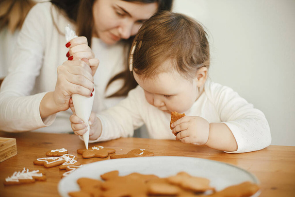 Aranyos kislány és anya együtt díszítő karácsonyi mézeskalács cookie-k cukormázas fa asztal a modern szobában. Anyuci lánya autentikus szép pillanatok. Üdülési előkészületek - Fotó, kép