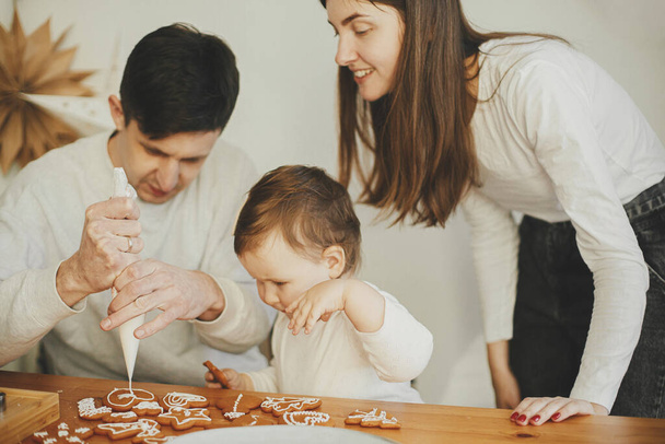 Stylish mother, father and cute little daughter decorating christmas gingerbread cookies with icing in modern room. Happy family time together, authentic lovely moments. Xmas holiday preparations - 写真・画像
