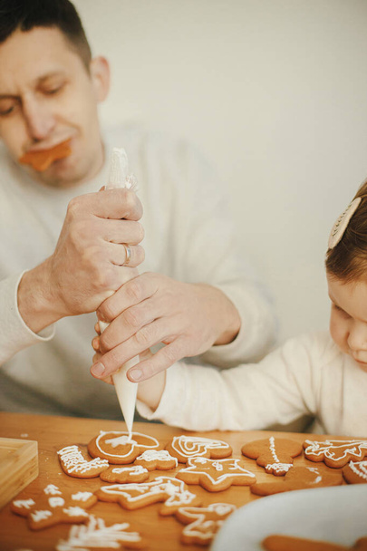 Happy stylish family decorating christmas gingerbread cookies with icing, funny authentic moments. Stylish dad and cute little daughter time together. Xmas holiday preparations - Photo, Image