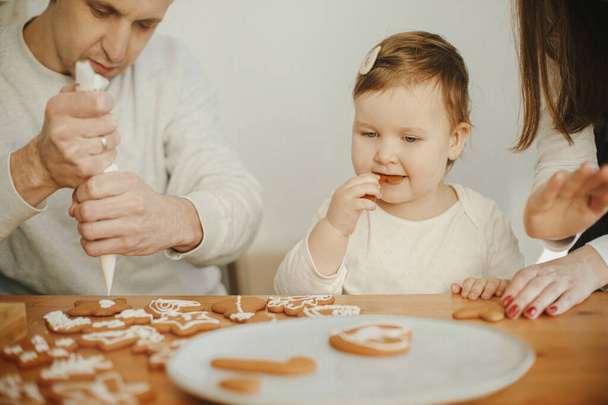 Stylish mother, father and cute little daughter decorating christmas gingerbread cookies with icing in modern room, funny authentic moments. Happy family time together. Xmas holiday preparations - Photo, Image