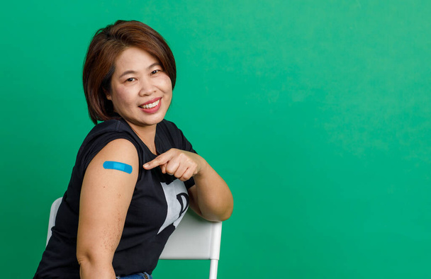 Estúdio tiro de mulher asiática de meia-idade paciente sentar sorriso e mostrar bandagem de gesso azul no braço depois de receber coronavírus covid 19 vacinação pelo médico na clínica em fundo verde. - Foto, Imagem
