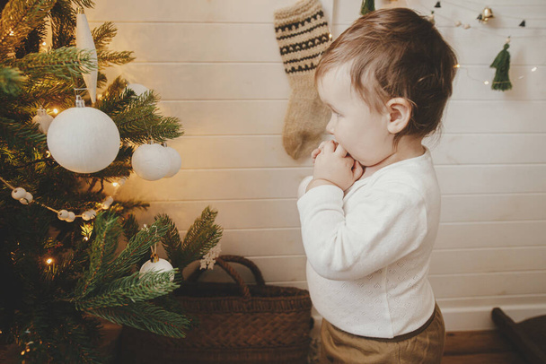 Cute baby girl patrząc na nowocześnie urządzone choinki z białymi bombkami. Szczęśliwy stylowy maluch dziewczynka stoi na choince w świątecznym pokoju skandynawskim. Autentyczne piękne chwile - Zdjęcie, obraz