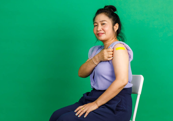Студийный снимок азиатской женщины средних лет сидеть улыбку и показать желтую гипсовую повязку на руку после получения коронавируса ковида 19 вакцинация врачом в клинике на зеленом фоне. - Фото, изображение