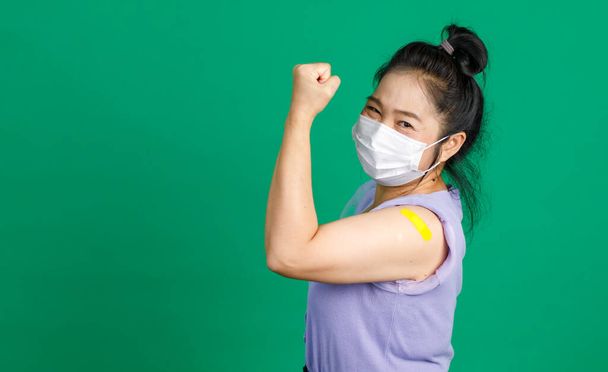 Studyjne ujęcie azjatyckiej szczęśliwej zdrowej pacjentki w średnim wieku nosi maskę na twarzy, patrzy na kamerę trzymającą pięść, pokazuje mięśnie i niebieski plaster bandażowy po otrzymaniu szczepionki przeciw koronawirusowi covid 19. - Zdjęcie, obraz