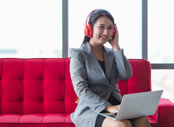 Marzycielska azjatycka przedsiębiorczyni w słuchawkach siedząca na kanapie w przedpokoju centrum biznesowego i ciesząca się piosenkami z laptopa notebooka z łatwą pozą podczas przerwy w pracy - Zdjęcie, obraz