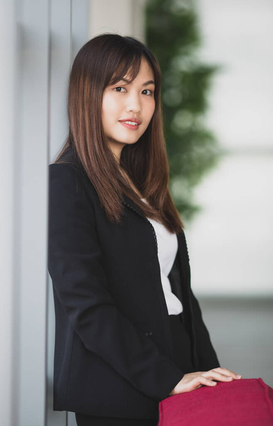 Πορτρέτο της χαριτωμένο και νεαρή Ασιάτισσα επιχειρηματίας ή γραμματέας σε κομψό ντύσιμο στέκεται με φιλικό χαμόγελο πρόσωπο δίπλα από τα παράθυρα στην αίθουσα γραφείων και κοιτάζοντας την κάμερα - Φωτογραφία, εικόνα