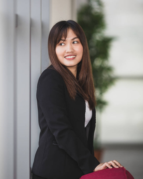 Porträt einer netten und jungen asiatischen Unternehmerin oder Sekretärin im eleganten Outfit, die mit freundlichem Lächeln vor den Fenstern im Büroflur steht und nach draußen blickt - Foto, Bild
