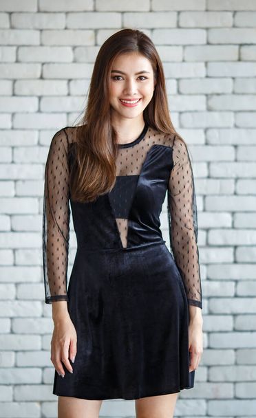 Портрет молодой и милой милой азиатки в черном платье, стоящей на фоне белой кирпичной стены с дружелюбной позой и положительным теплым оттенком. - Фото, изображение