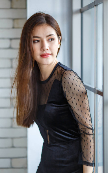 Вид сбоку портрет очаровательной азиатской женщины, лаосской этнической принадлежности, в элегантном черном платье, стоящем с симпатичной и спокойной позой у окна и глядя в сторону - Фото, изображение