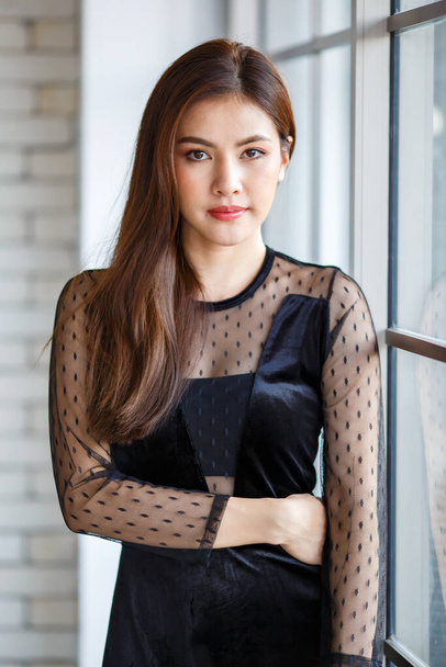 Portret halve lengte van schattige charmante brunette Aziatische vrouw, Lao etniciteit, in stijlvolle zwarte jurk staan en poseren met vriendelijke positieve en zelfverzekerde pose in de buurt van glas raam. - Foto, afbeelding