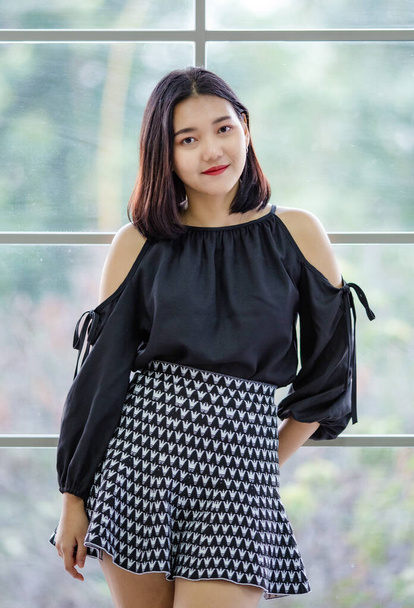 Portrait half lenght of cute charming brunette Asian female in stylish black dress standing and pose with friendly positive and self confident pose near glass window. - Photo, Image