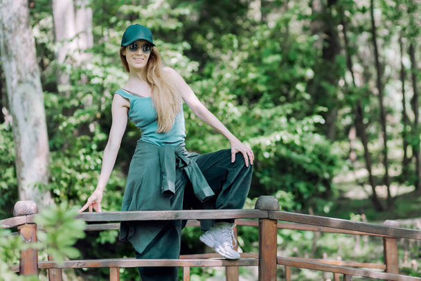 Сучасна блондинка в хіп-хоп одязі стоїть з піднятою ногою на дерев'яному барі мостових перил в зеленому парку
 - Фото, зображення