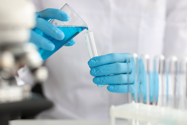 Wetenschapper met handschoenen giet blauwe vloeistof in reageerbuis in laboratorium - Foto, afbeelding